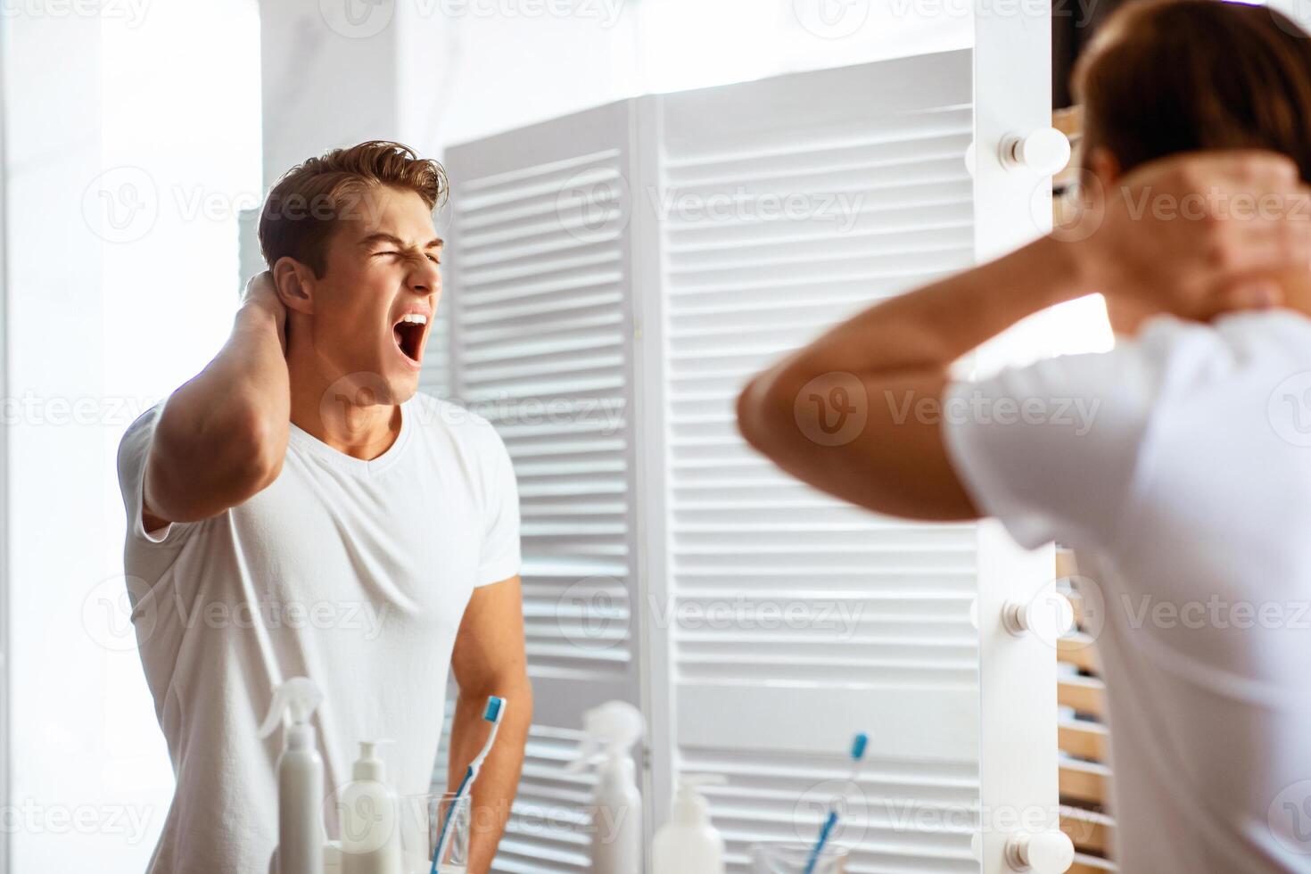 Portrait of sleepy young guy yawning, standing in front of a mirror in bathroom in the morning, view over the shoulder photo