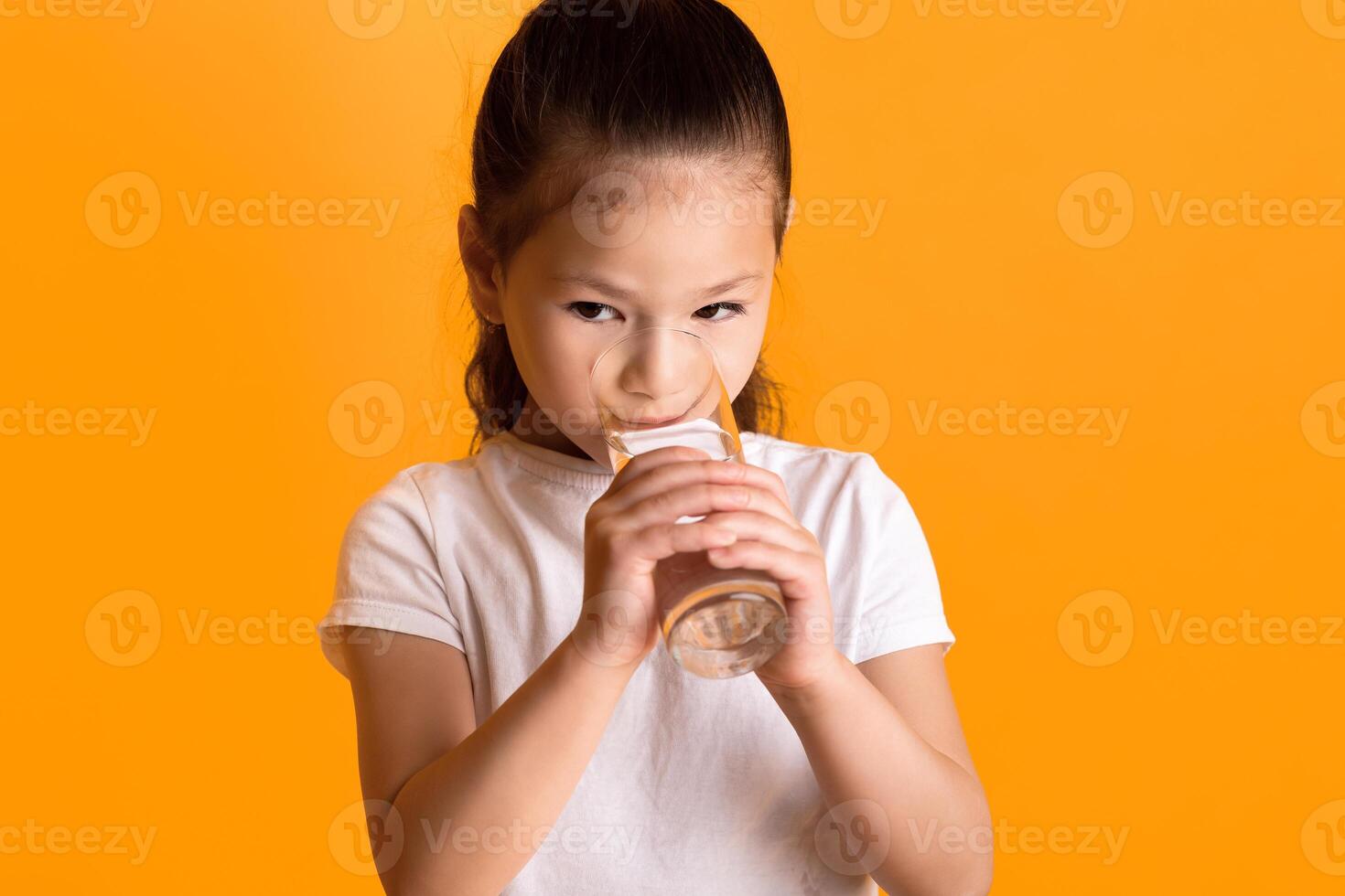 Hydration Concept. Little adorable asian girl drinking spring water at studio, looking away. Closeup portrait of beautiful kid holding glass of mineral water, hydrating her body in the morning photo