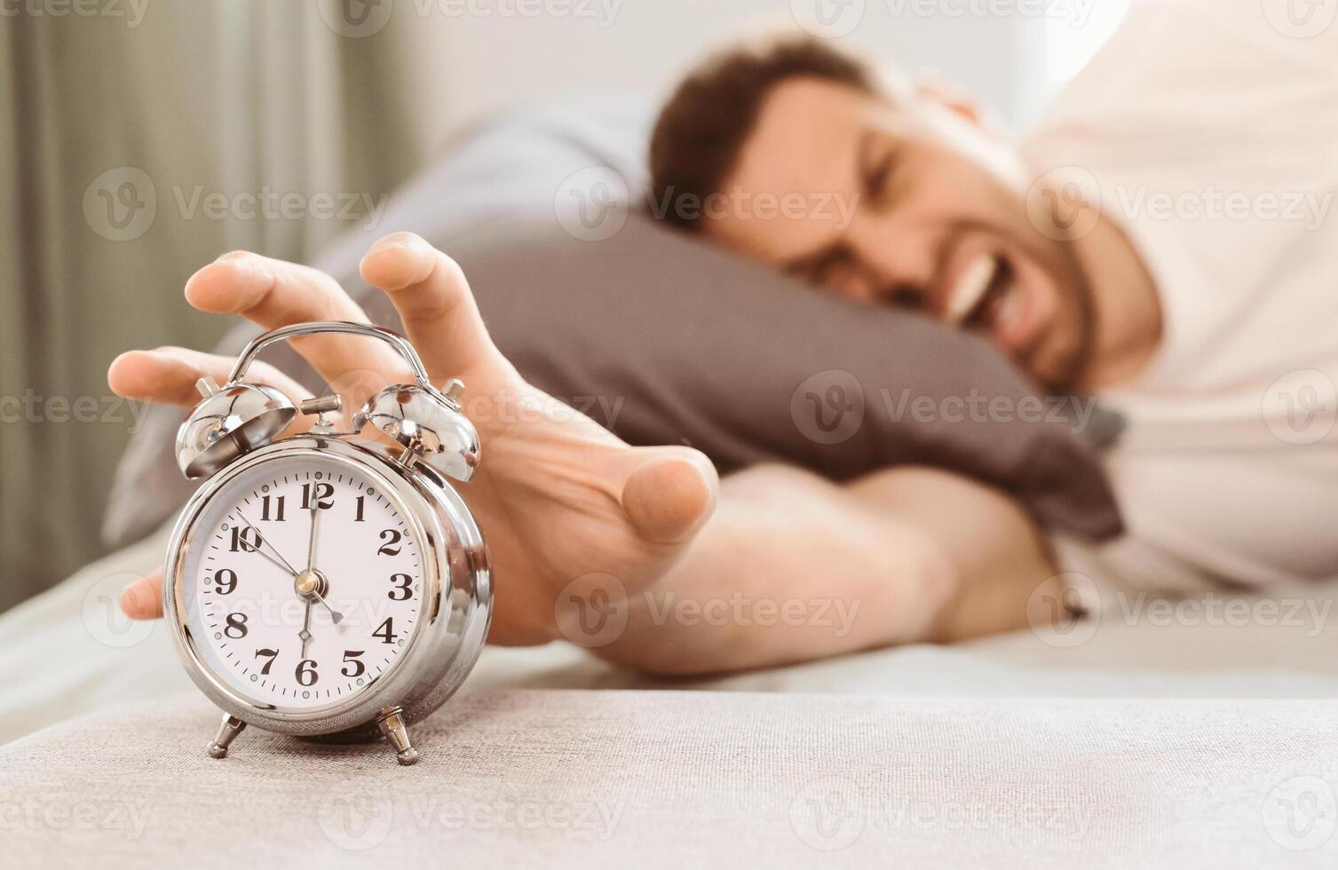 Bad Morning. Angry Man Turning Off Alarm-Clock Waking Up Early Lying In Bed At Home. Selective Focus, Shallow Depth photo