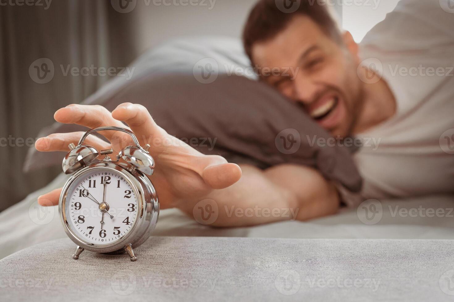 Angry Man Turning Off Alarm-Clock Shouting Waking Up Early In The Morning Lying In Bed At Home, Focus On Clock. Lack Of Sleep, Insomnia And Sleeplessness, Difficult Awakening. Low Light, Shallow Depth photo