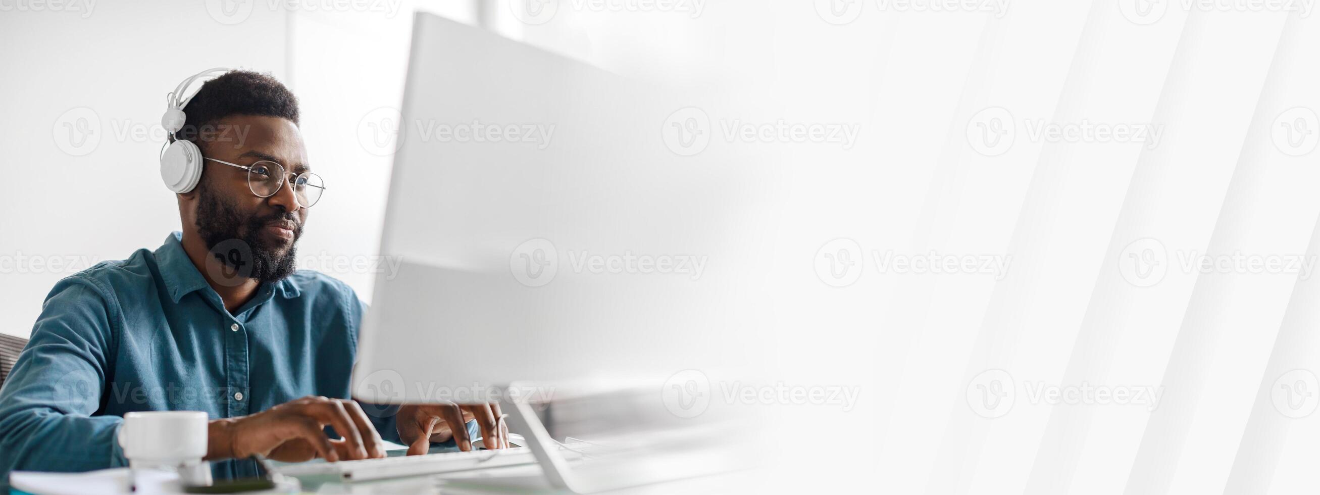 A young African American man works diligently at his desk in a modern office. He wears headphones and is engaged in tasks, showcasing a successful lifestyle as a manager and entrepreneur, copy space photo