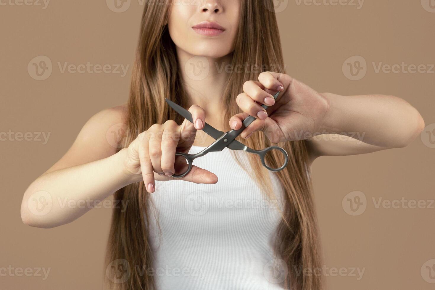 Beautiful millennial woman with perfect long hair trying to break scissors on brown background, selective focus. Lovely young lady unwilling to do haircut, having strong and healthy locks photo