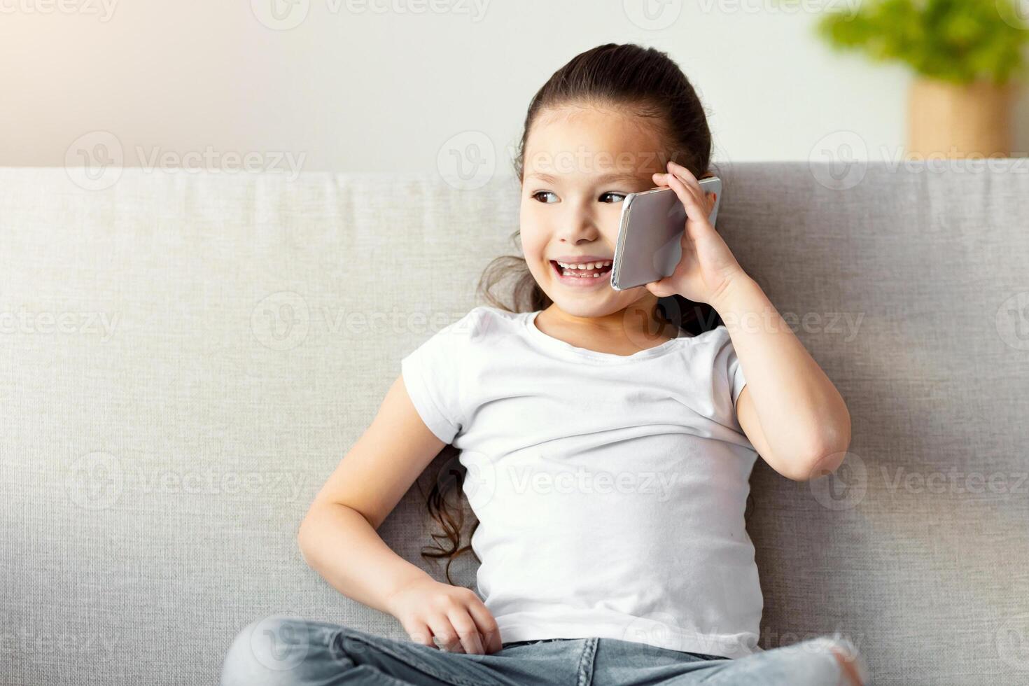 Little Chinese Girl Talking On Mobile Phone Chatting With Friend Sitting On Couch At Home. Child And Gadget. Free Space photo