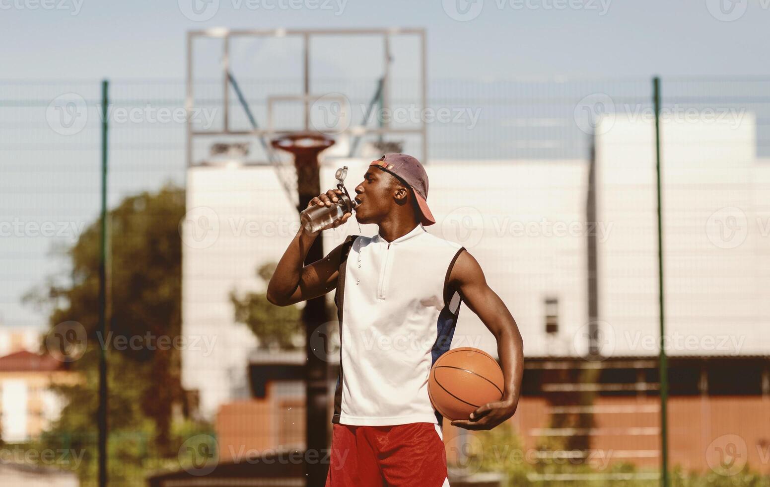 Young black basketball player drinking water to refresh himself after training on outdoor court, panorama photo