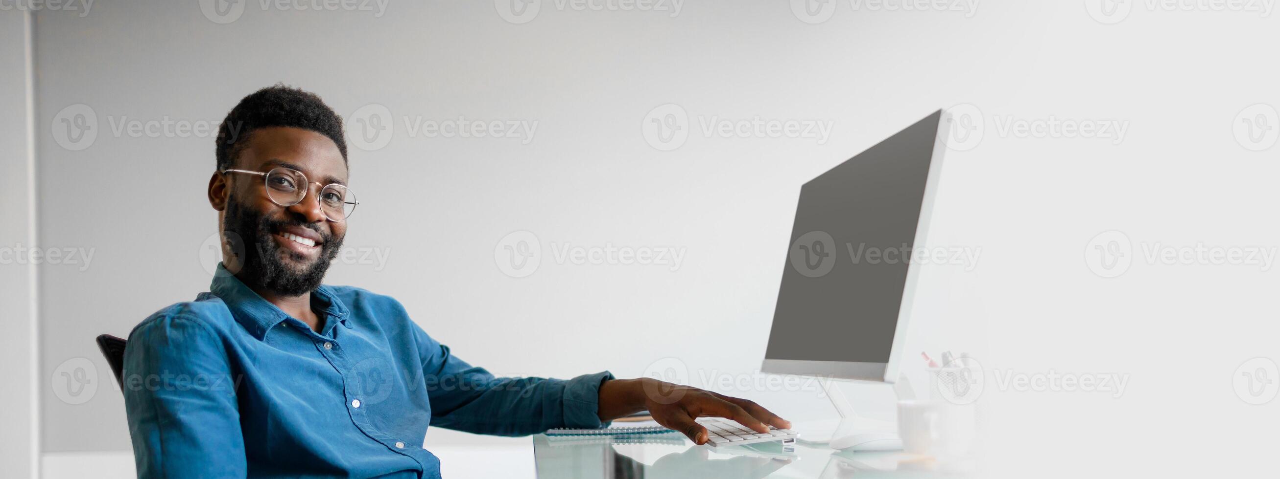 A young African American entrepreneur sits at his office desk, feeling accomplished and engaged in his work. Bright and modern environment enhances his professional lifestyle, copy space photo