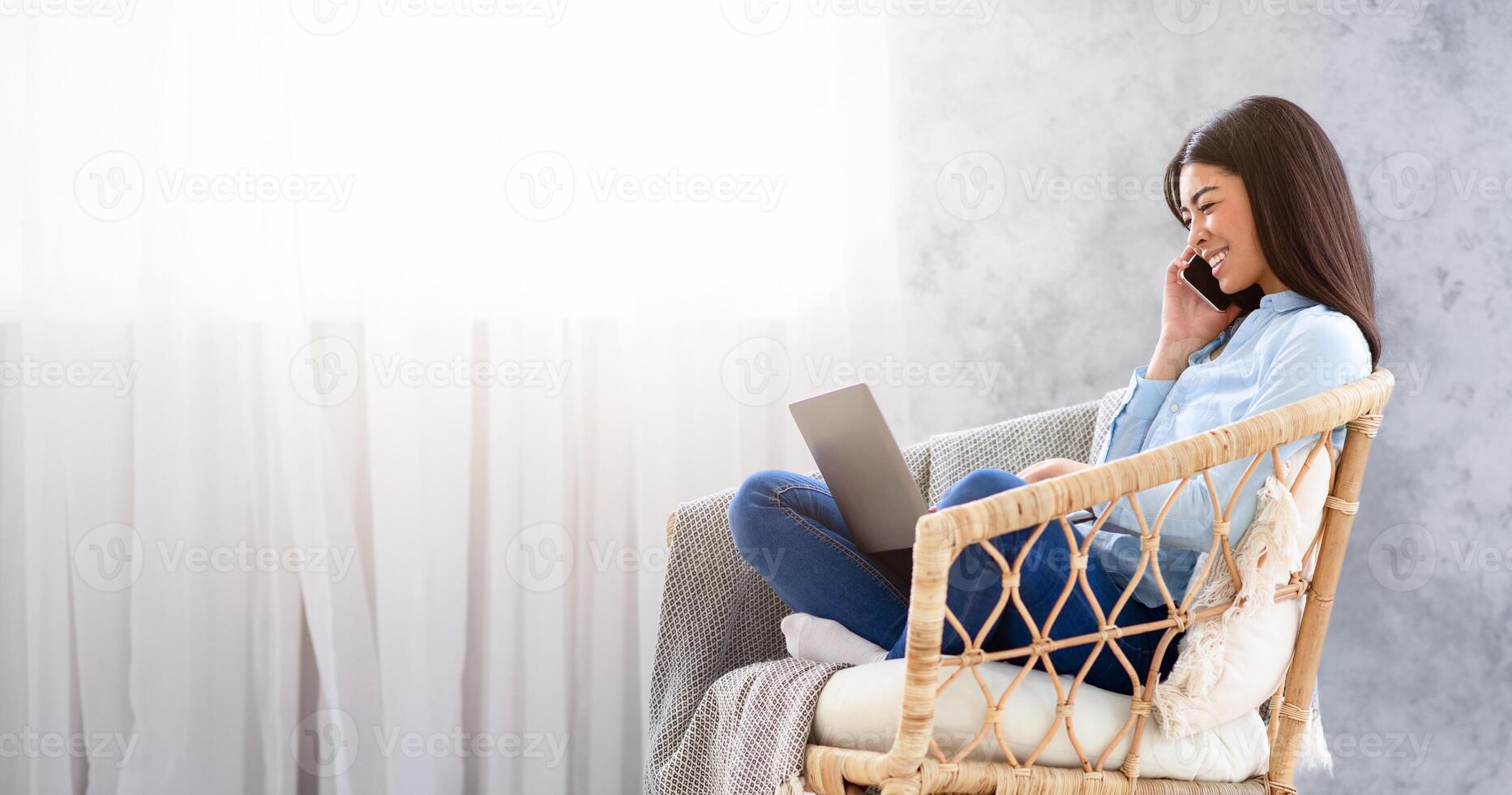 Young Asian Woman Working On Laptop And Talking On Cellhone While Sitting In Stylish Wicker Chair At Home, Side View With Copy Space photo
