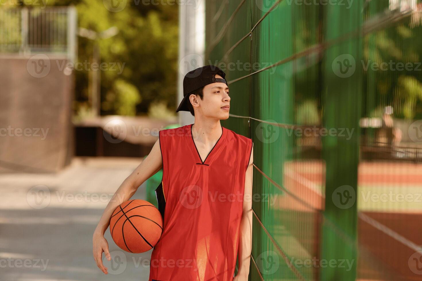 Thoughtful young Asian basketball player with ball on outdoor court, copy space. Millennial sportsman resting after game, training for tournament outside. Professional sports concept photo