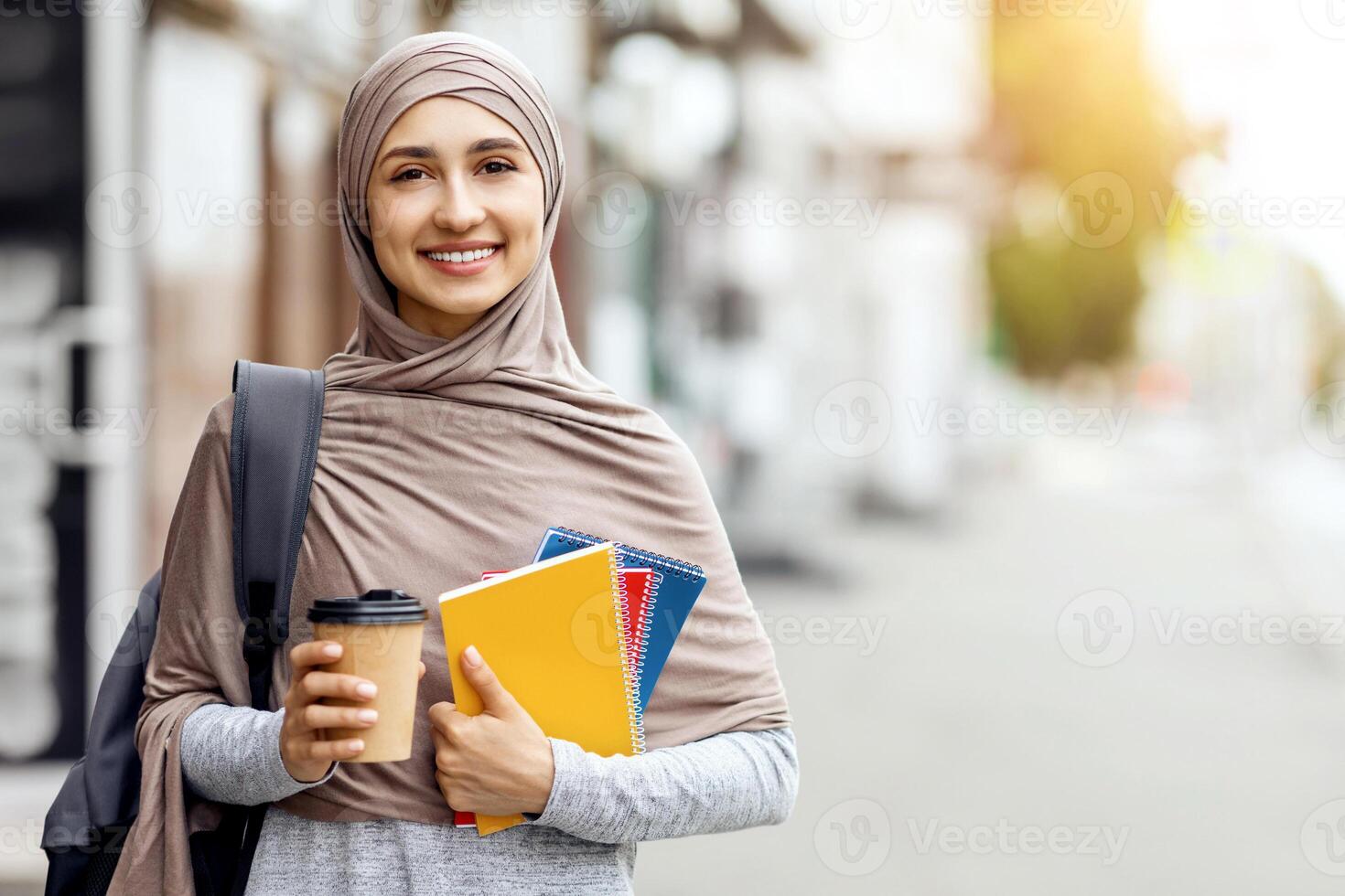 Cheerful girl in hijab having morning coffee on the street before university, free space photo