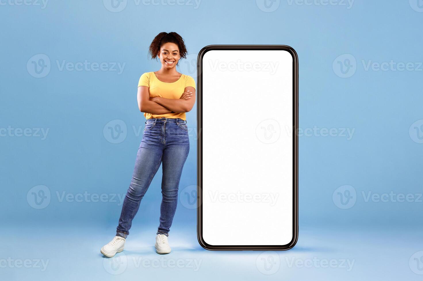 Beautiful African American lady standing near huge smartphone with empty white screen on blue studio background, space for mockup. Newest mobile application, website, advertisement concept photo