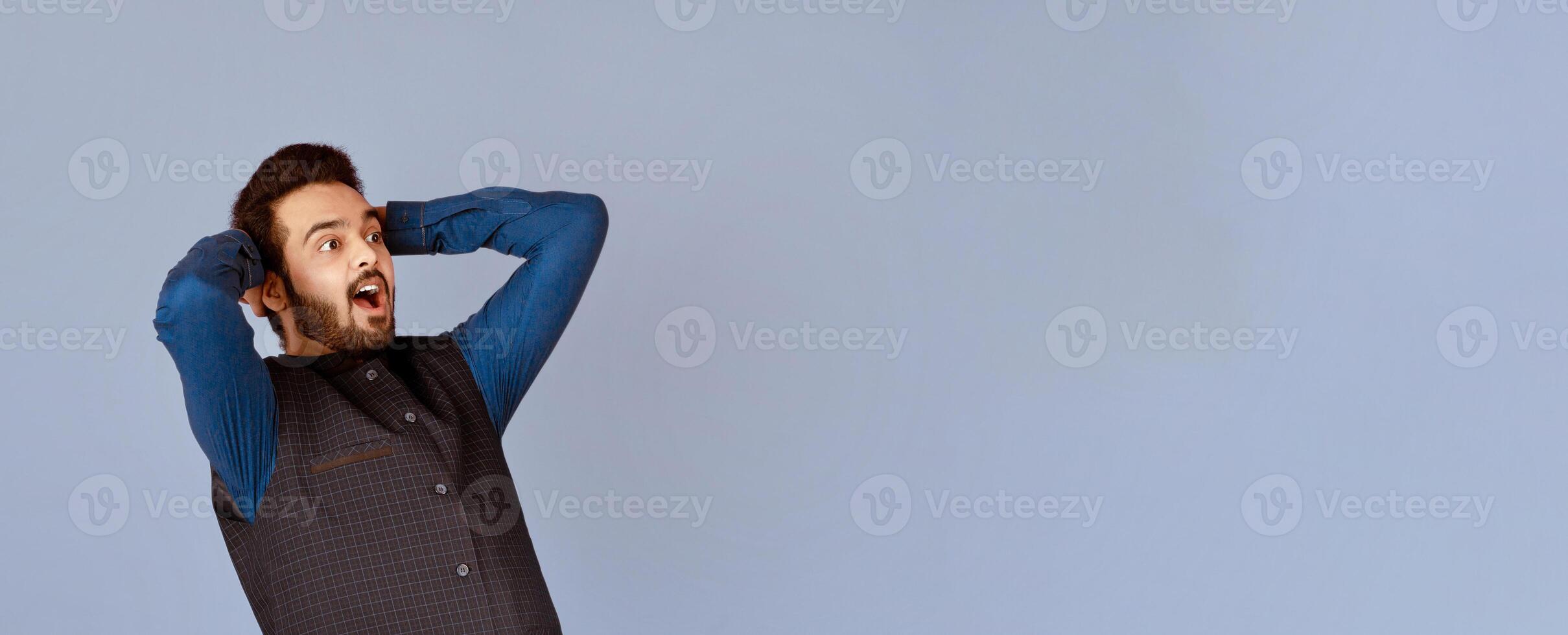 Shocked Indian guy holding his head and screaming WOW on violet studio background, panorama photo