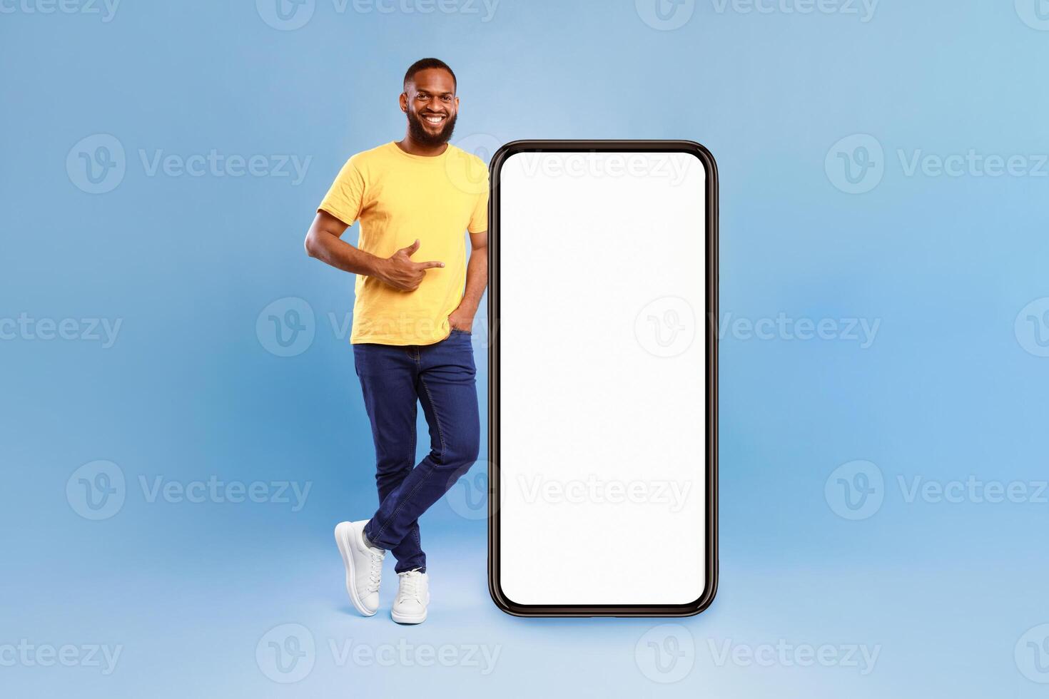 Positive African American guy pointing at big smartphone with mockup for mobile app, recommending cool website over blue studio background. Cellphone screen with space for your advertisement photo