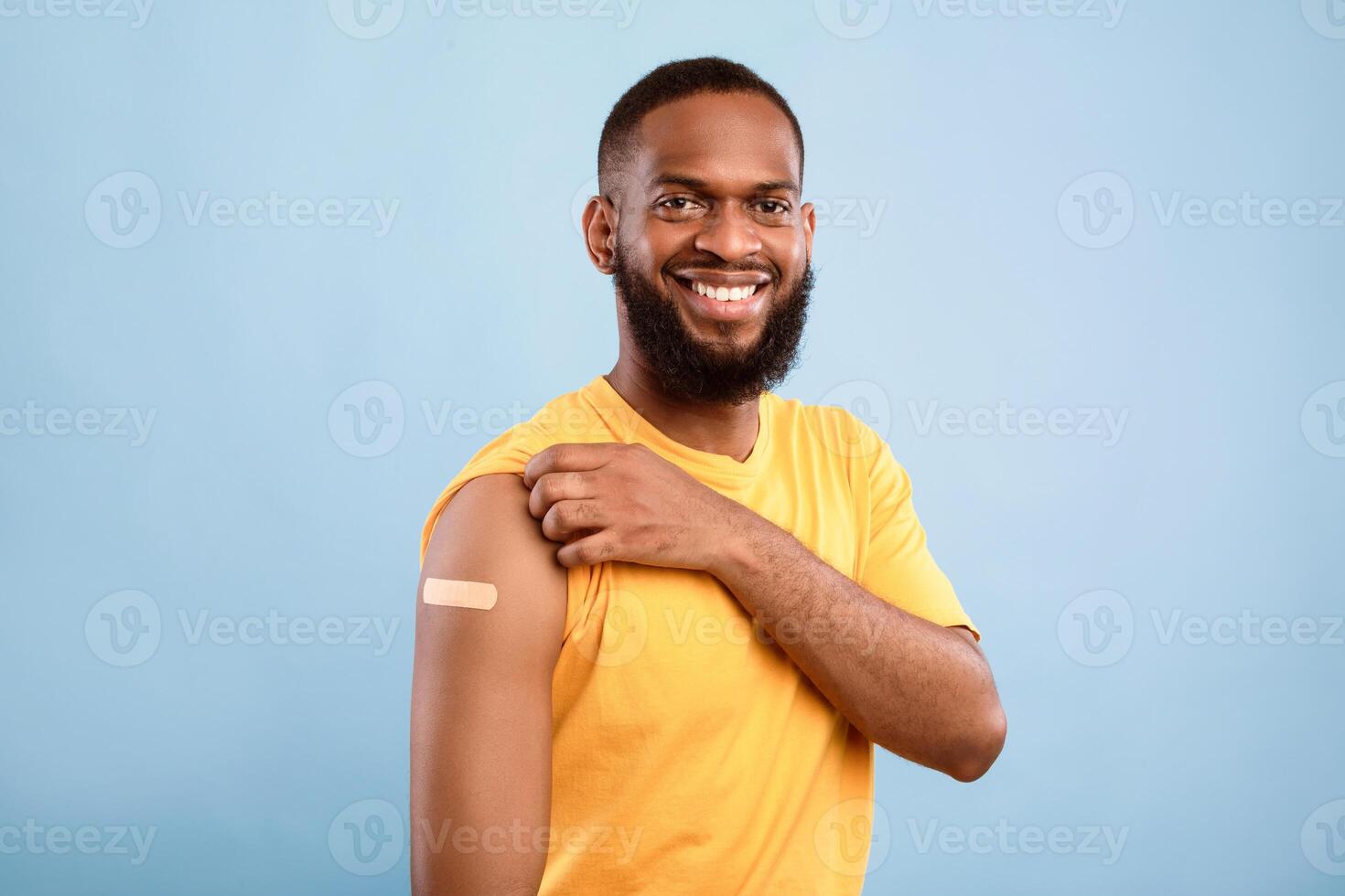 Happy African American guy smiling at camera after flu vaccine injection, showing arm with band aid after flu immunization on blue studio background. Stop flu vaccination campaign photo