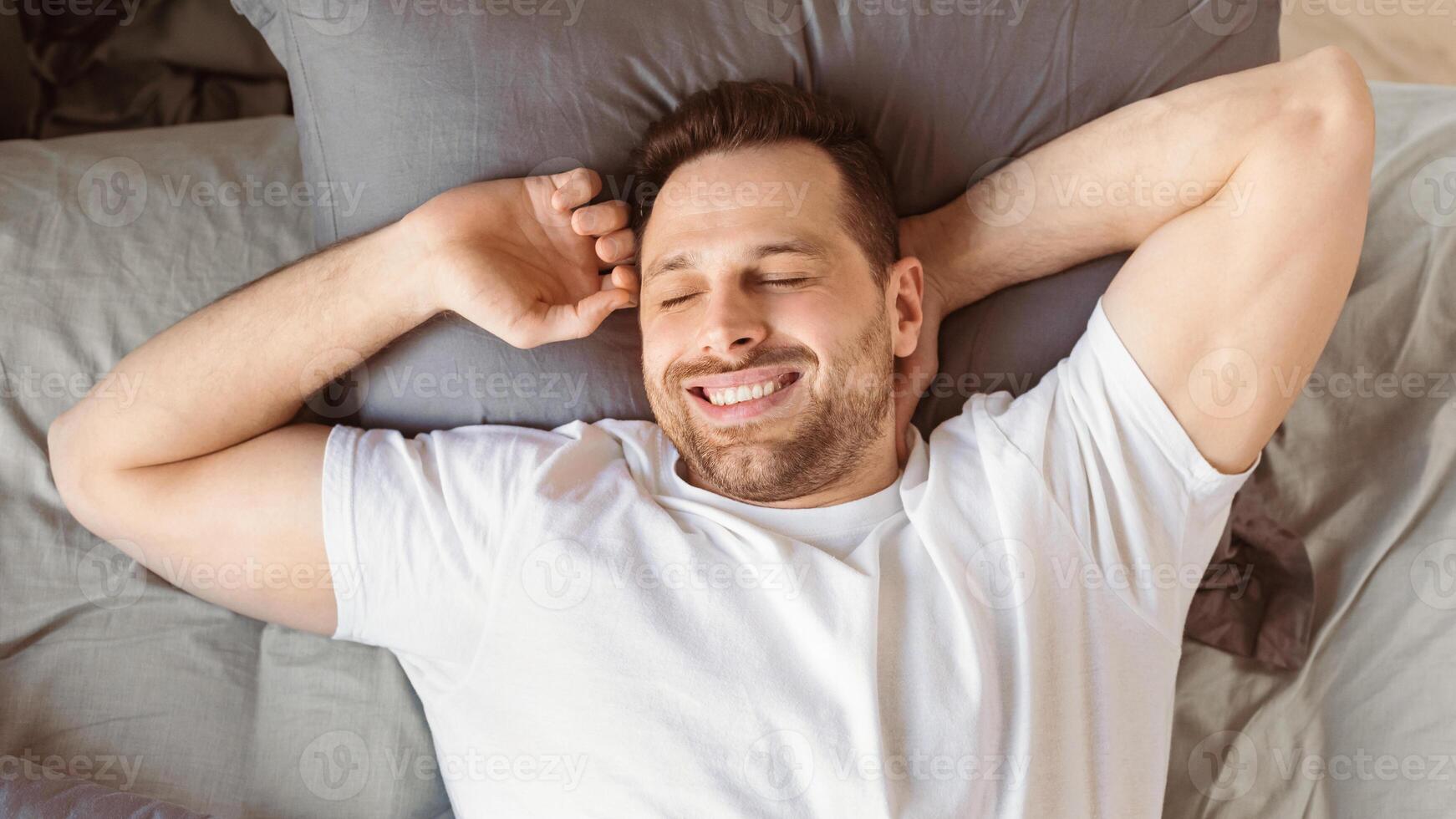 Good Morning. Joyful Guy Waking Up Yawning And Stretching Hands Lying In Comfortable Bed In Bedroom At Home, Smiling With Eyes Closed. Healthy Male Sleep. View From Above, Panorama photo