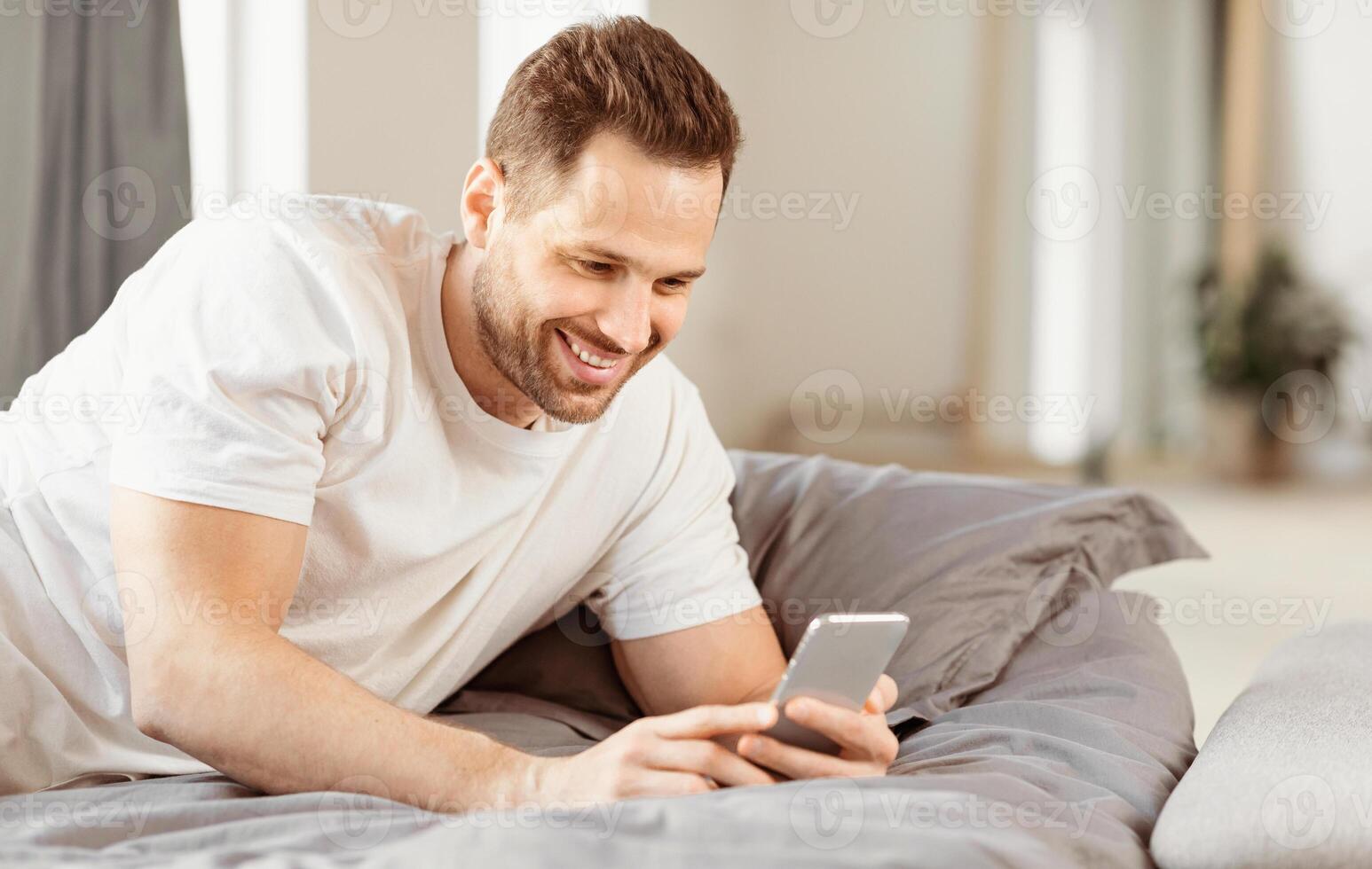 Mobile App. Cheerful Guy Using Smartphone Lying In Bed In Bedroom At Home. Man Browsing Internet On Phone. Application For Cellphone Advertisement. Free Space For Text photo
