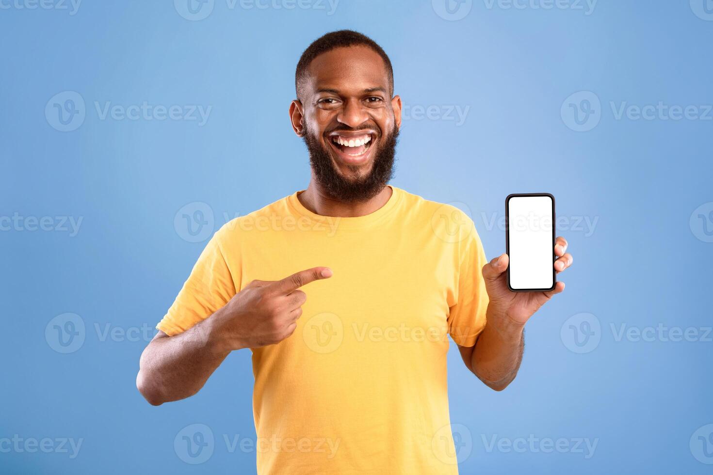 Check out this mobile app. Excited African American man presenting smartphone with blank screen, offering space for website or ad, smiling at camera on blue studio background, mockup photo
