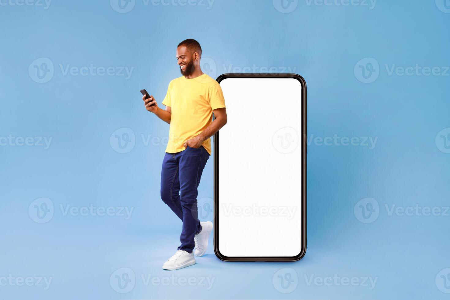 Handsome young African American man using mobile phone while standing near huge smartphone with empty screen over blue studio background. Mockup for website or application, space for advertisement photo