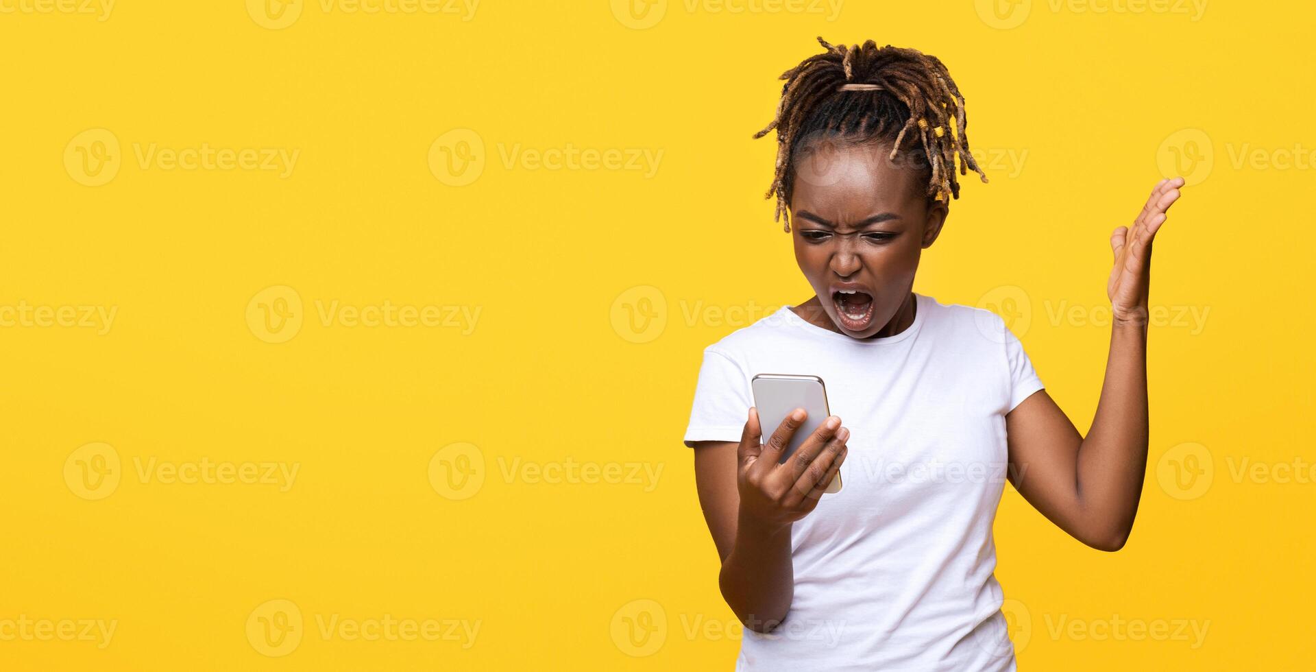 Furious black lady standing on yellow studio background, screaming at smartphone in her hand, gesturing, expressing anger, panorama with empty space. Unhappy african american lady holding mobile phone photo