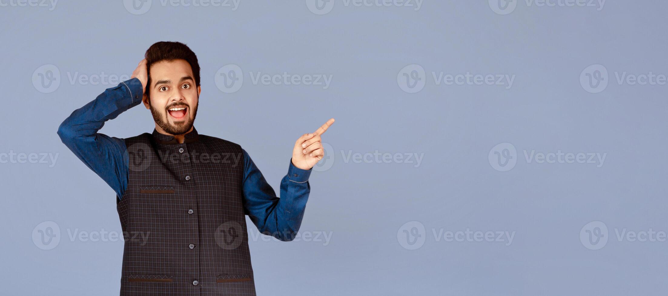 Excited Indian guy holding his head in disbelief and pointing at empty space over violet background photo