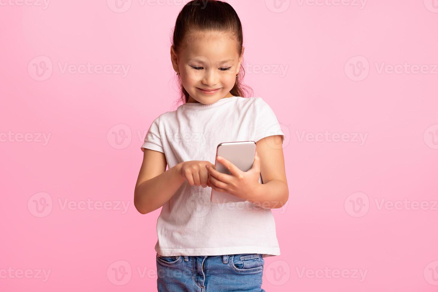 Kids And Technology. Smiling asian child holding and using cellphone, sending message to parents or friends, browsing internet, watching or cartoons, standing over pastel pink wall, copy space photo