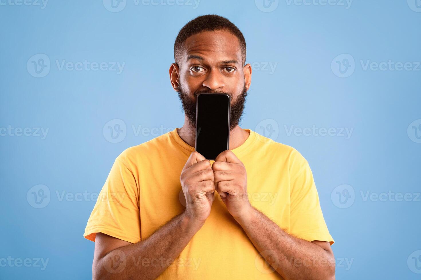 Handsome African American man covering is mouth with blank cellphone with mockup for app or website design over blue studio background. Cool guy showing mobile advertisement photo