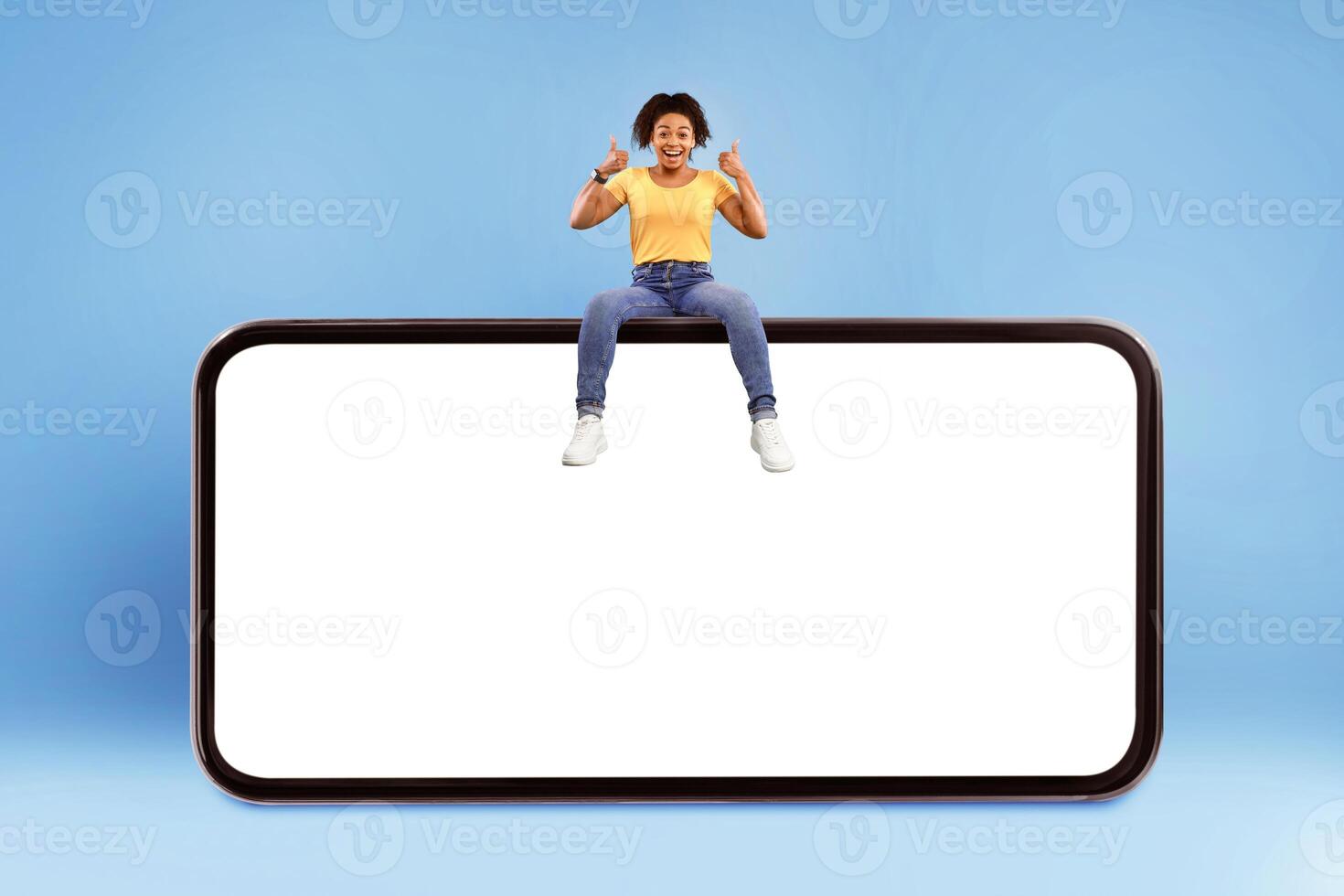Positive millennial African American woman showing thumb up gesture, sitting on top of huge smartphone over blue studio background, empty space for mobile app on screen. Website or advert mockup photo