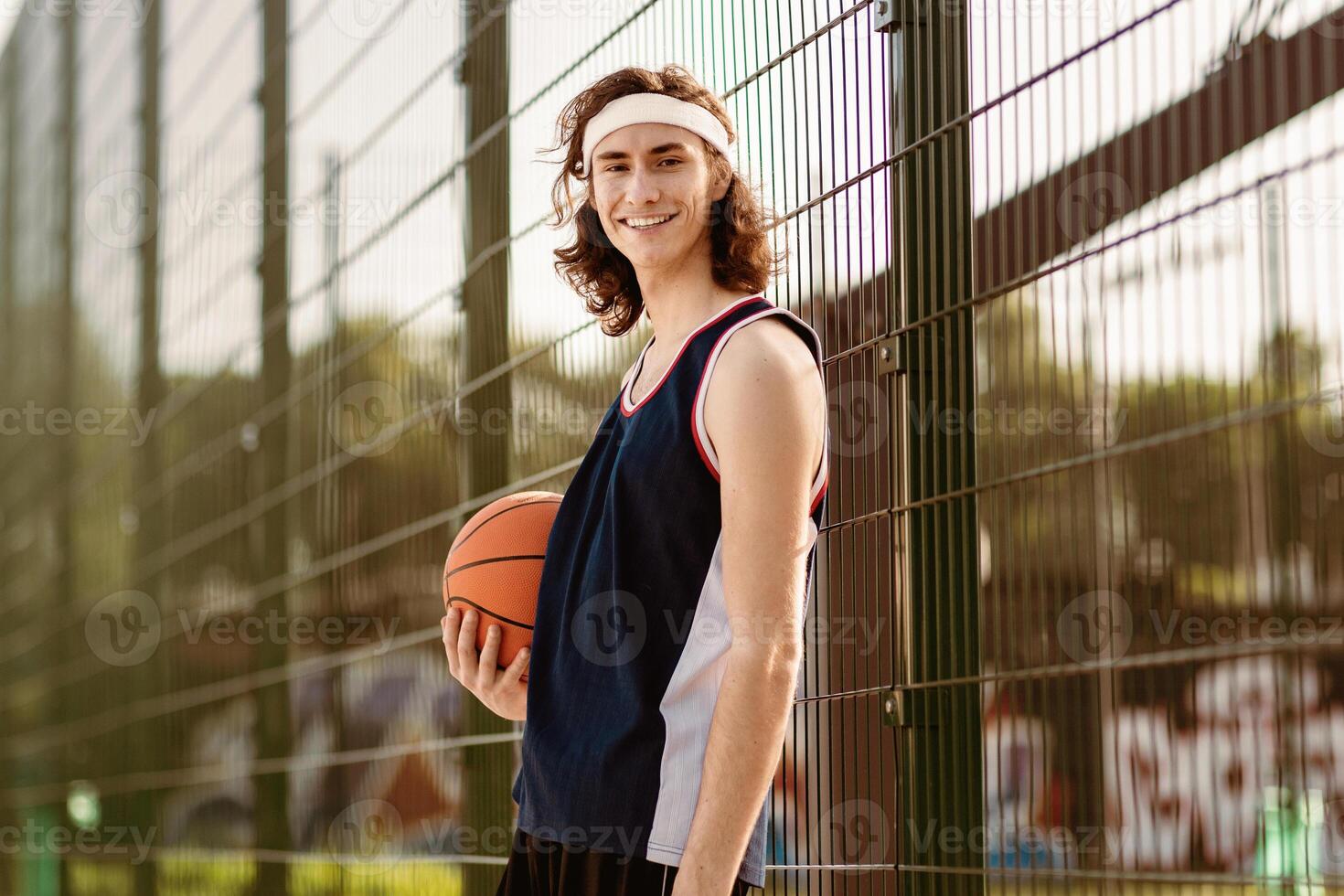 Portrait of positive millennial basketball player in sports uniform posing at outdoor stadium before game, empty space. Young basketballer smiling at camera after training for big competition photo