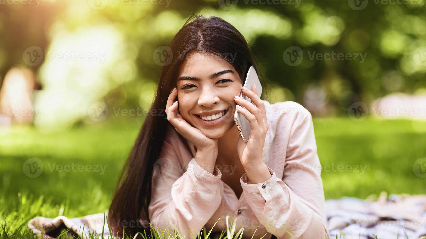 Pleasant Call. Happy Korean Girl Talking On Mobile Phone While Relaxing On Plaid In Park, Lying On Lawn And Speaking On Cellphone, Smiling At Camera photo