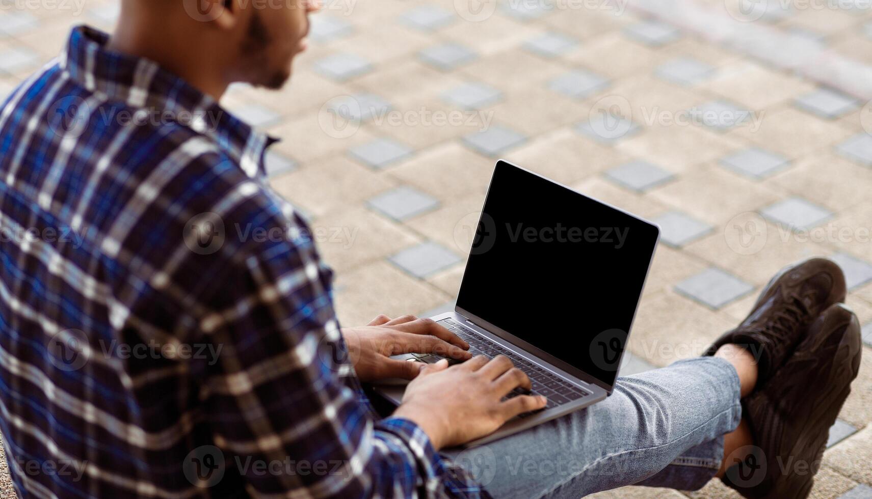 Technology Concept. View over the shoulder of african guy using pc with black blank screen outdoors, mock up photo