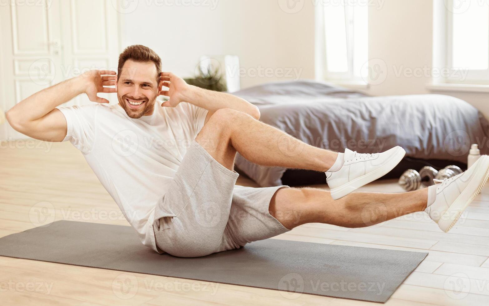 Home Training. Sporty Man Doing Knee-To-Elbow Crunches Abs Exercise On Gymnastics Mat Indoors In The Morning, Smiling To Camera. Male Fitness Exercises And Workout Routine Concept photo