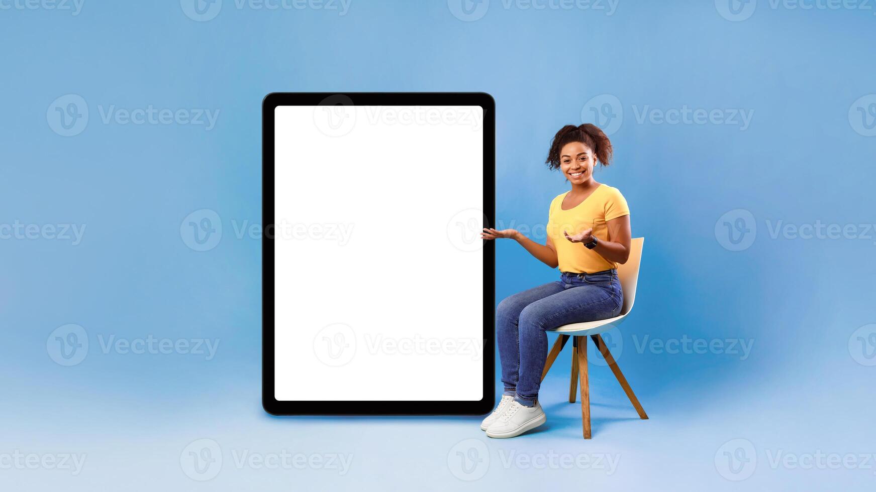 Happy millennial African American lady sitting on chair, pointing at giant tablet pc with white screen, offering empty space for app or website, presenting mockup for ad design, blue background photo