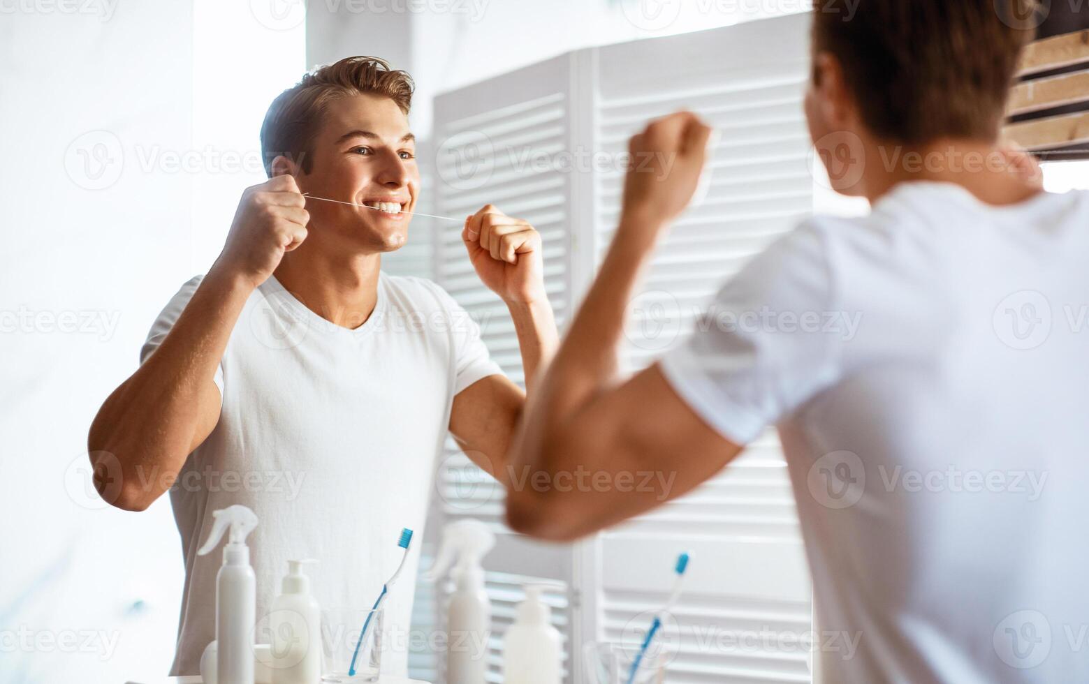 Oral Hygiene Concept. Portrait of handsome man using dental floss looking in the mirror at his reflection, view over the shoulder. Happy guy taking care of his strong teeth, doing morning routine photo