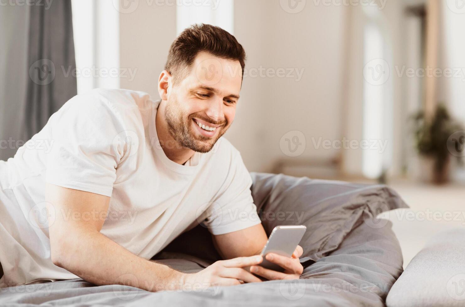 Happy Man Using Smartphone Lying In Bed At Home. Mobile Application. Copy Space photo
