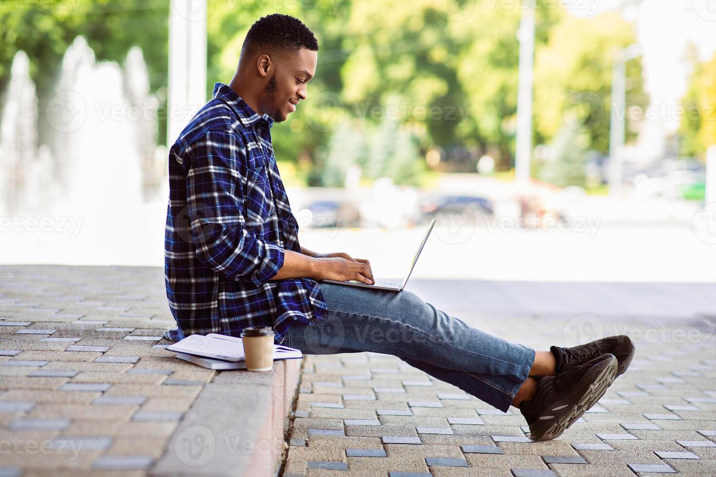 Side view of african guy working on laptop sitting on the border in downrown, selective focus photo
