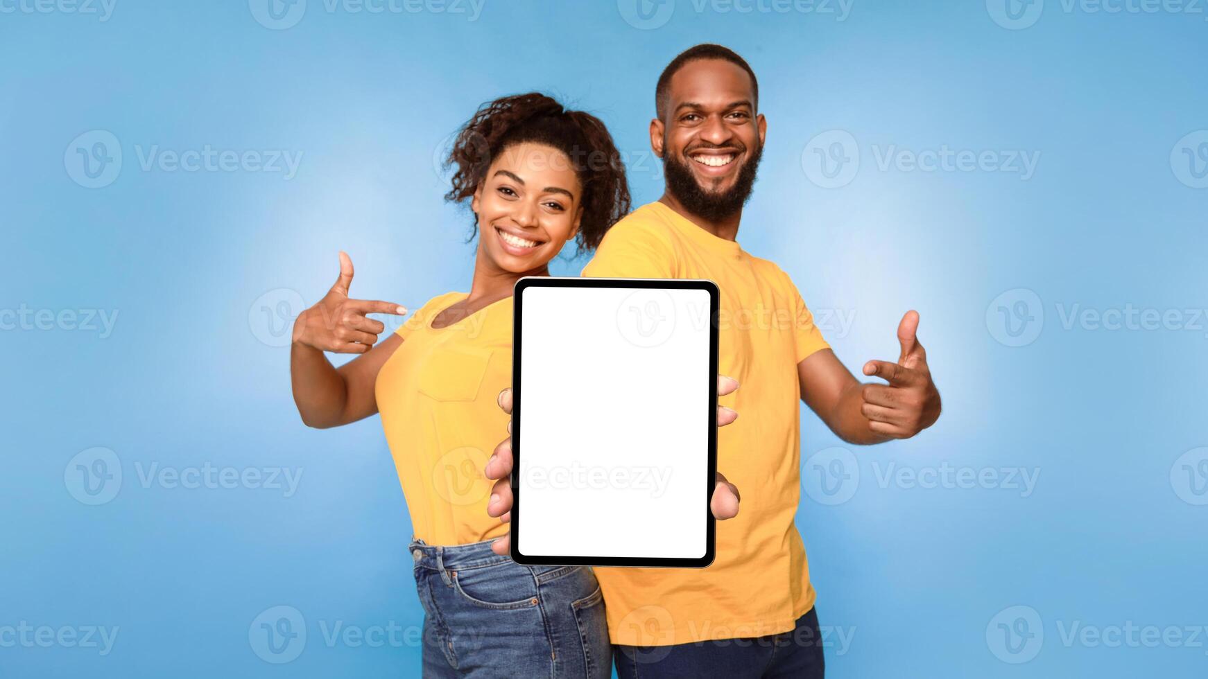 Cheerful African American couple pointing at digital tablet with blank white screen, recommending new application on blue studio background, mockup for design. Website, ad template photo