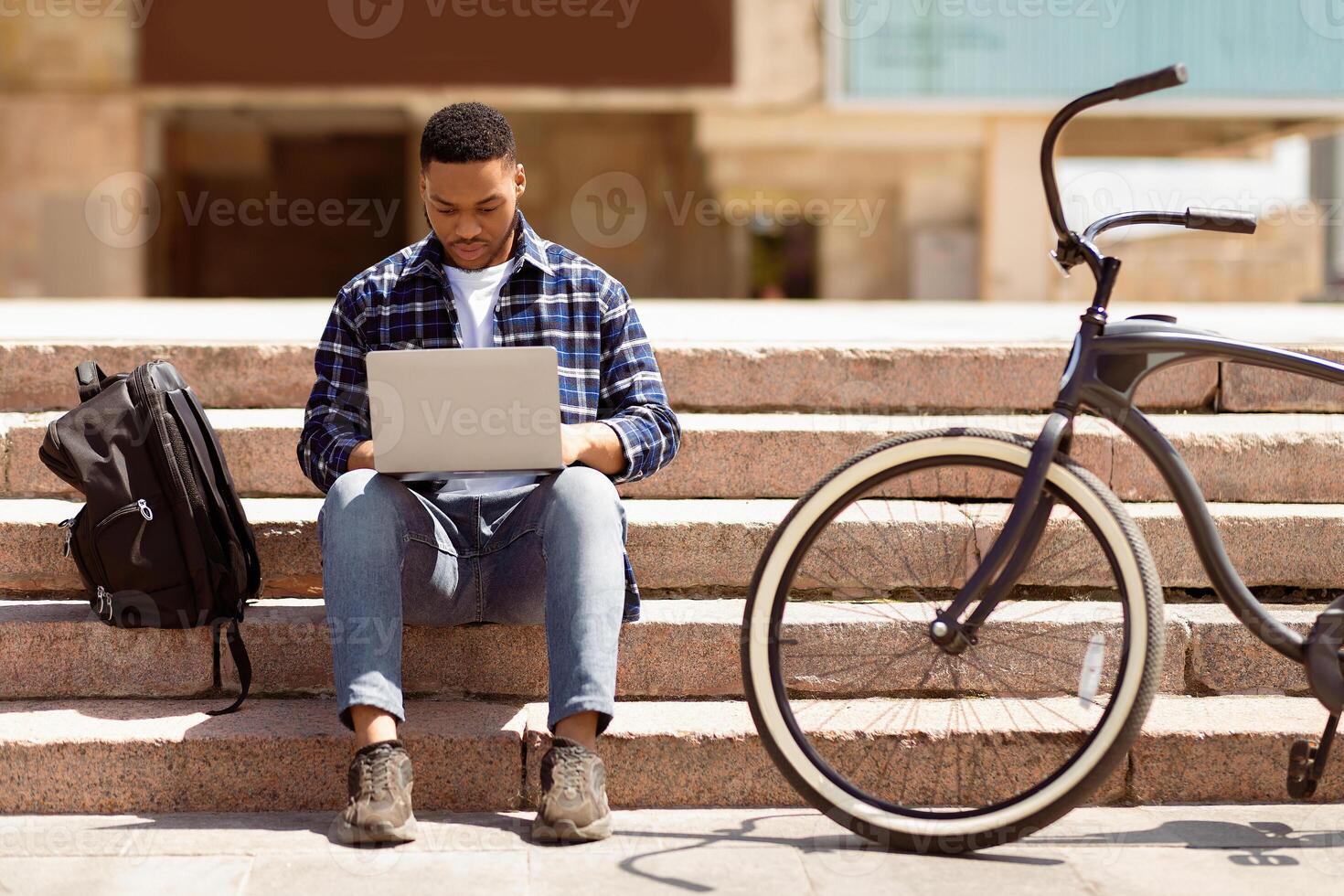 Front view of african guy working on laptop sitting on the steps in downtown, selective focus photo