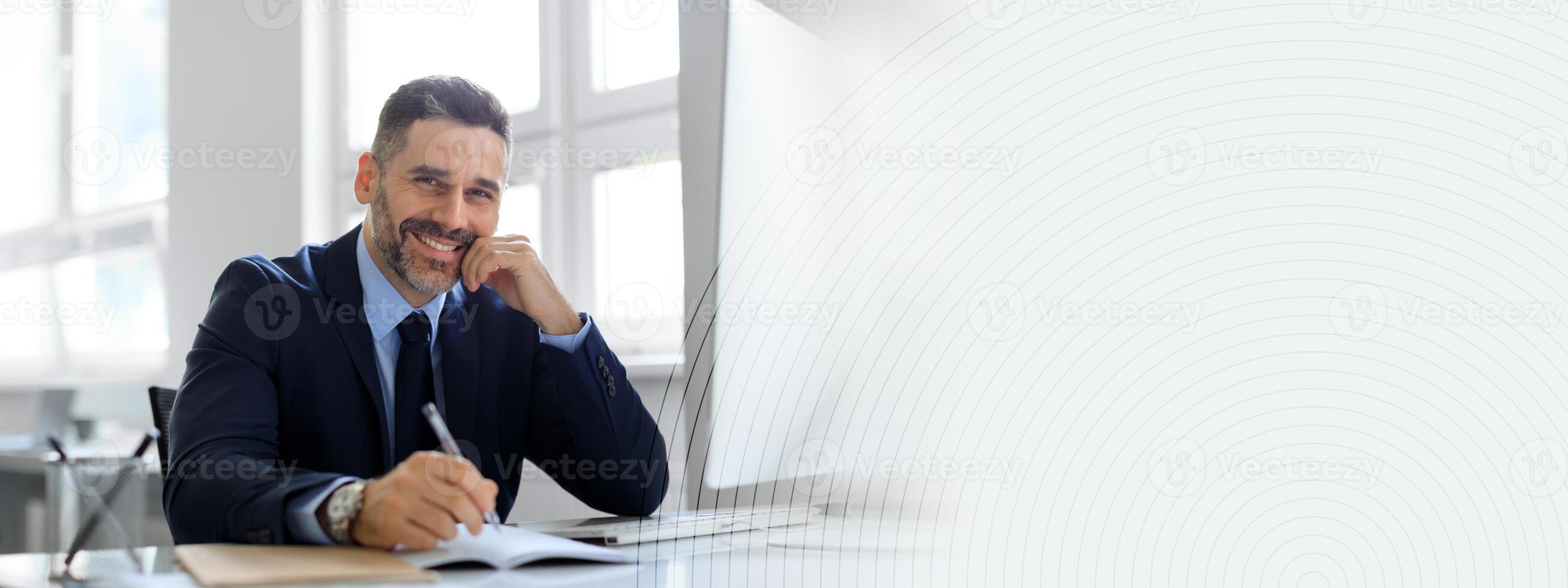 A middle aged Caucasian man in business attire is writing in a notebook while smiling. He appears focused and engaged in his work at a contemporary office desk, copy space photo