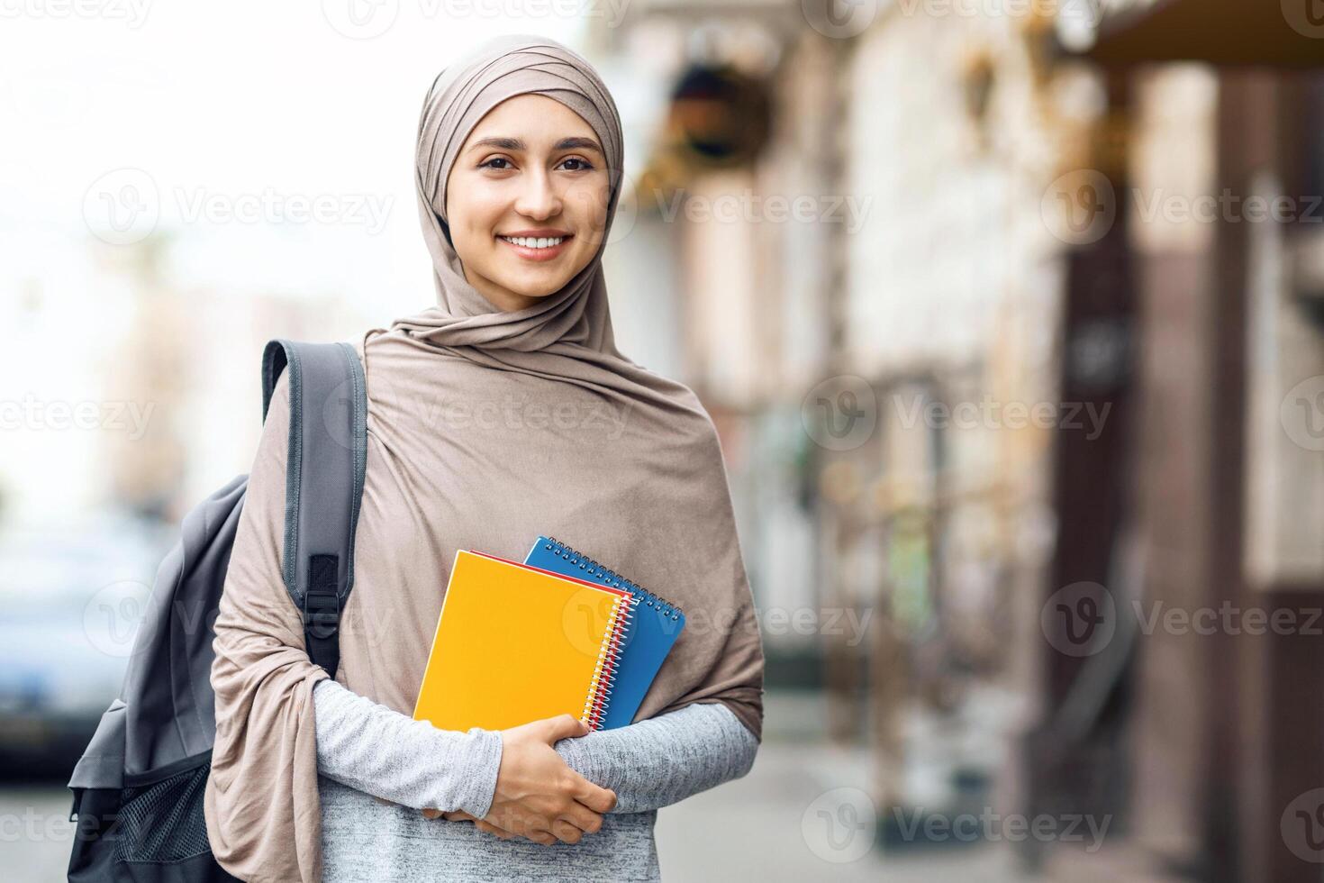 Muslim girl going to university, holding notepads and smiling at camera on the street, educational concept photo