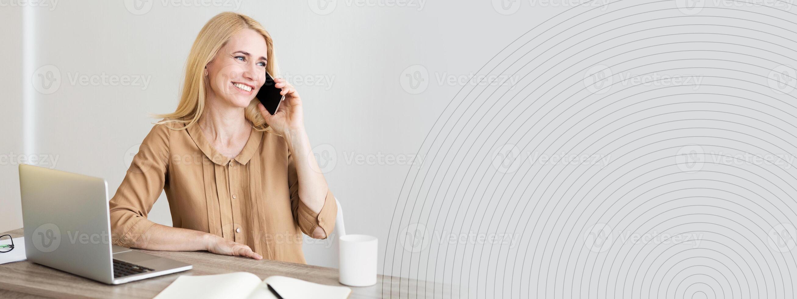 A woman engages in a phone conversation while sitting at a contemporary desk with a laptop and an open notebook. The workspace is well lit, creating a pleasant atmosphere for productivity, copy space photo