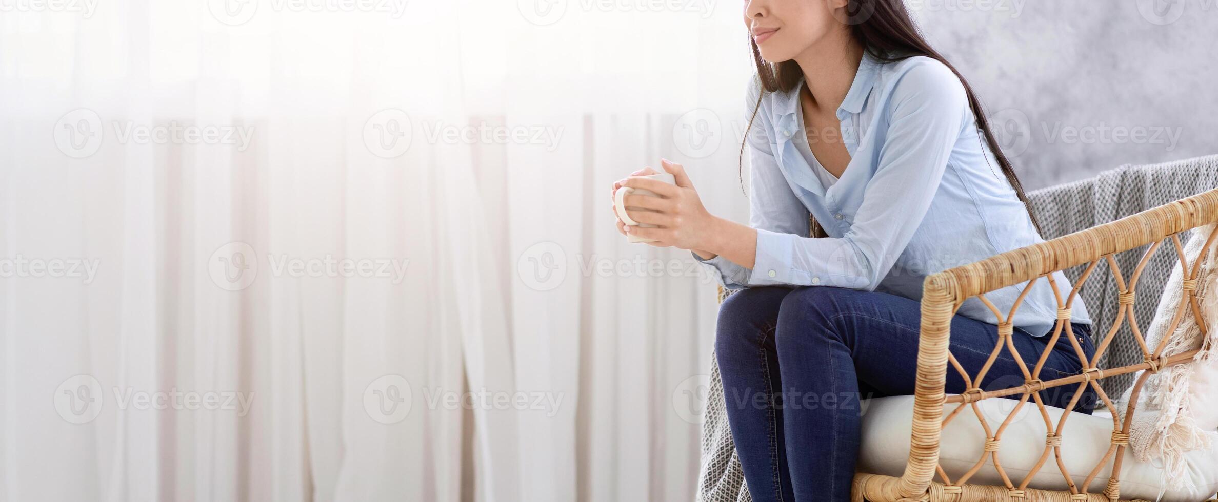 Domestic Relax. Smiling Korean Lady Resting In Armchair With Coffee At Home, Sitting In Wicker Chair Near Window With Cup Of Tea And Looking Away, Enjoying Weekend, Side View With Copy Space photo