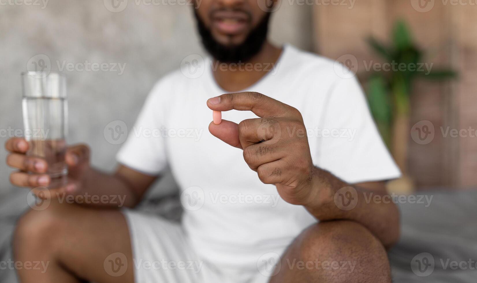 Closeup of young black man sitting on bed with glass of water, taking sleeping pills or painkillers at home. Unrecognizable millennial male using medications, holding tablet in hand, selective focus photo