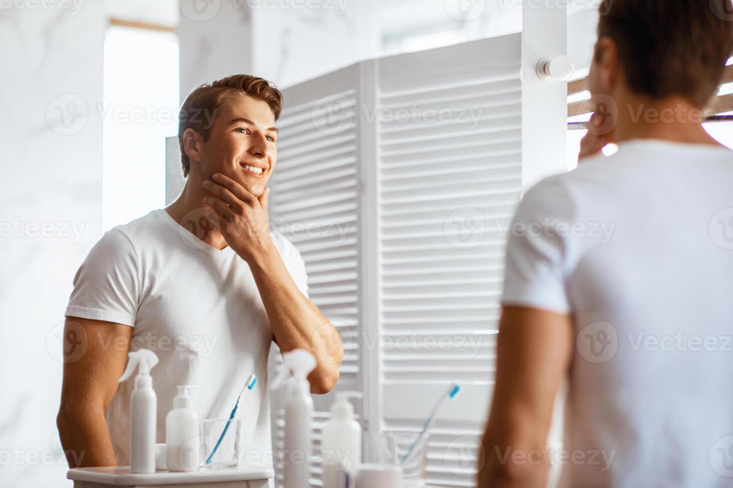 Young man touching his face with hand and gazing confidently while standing in front of the mirror photo