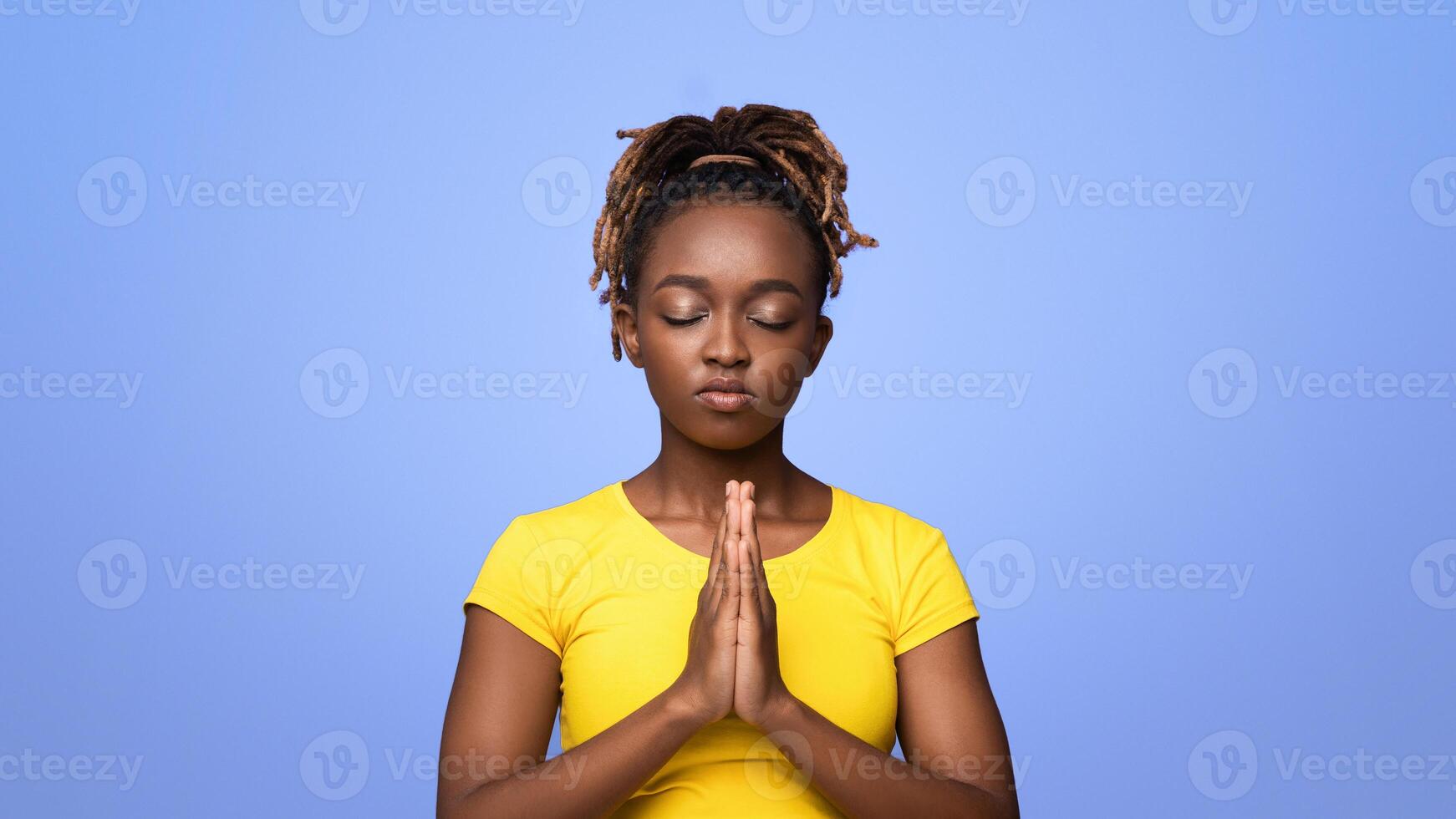Young black woman showing clasped hands in pleading gesture, pretty please on purple studio background, copy space. Religious african lady praying with closed eyes, having hope for better future photo