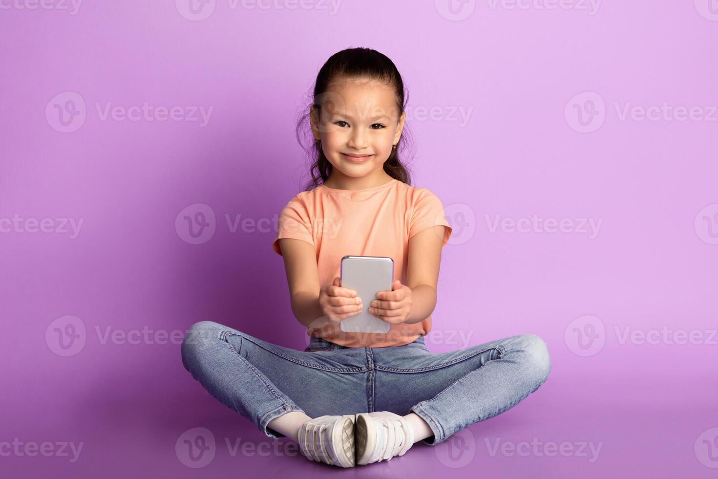 Childhood And Technology. Smiling asian girl holding and using smartphone, browsing internet, watching or cartoons, sitting on the floor over pastel purple background , copy space photo