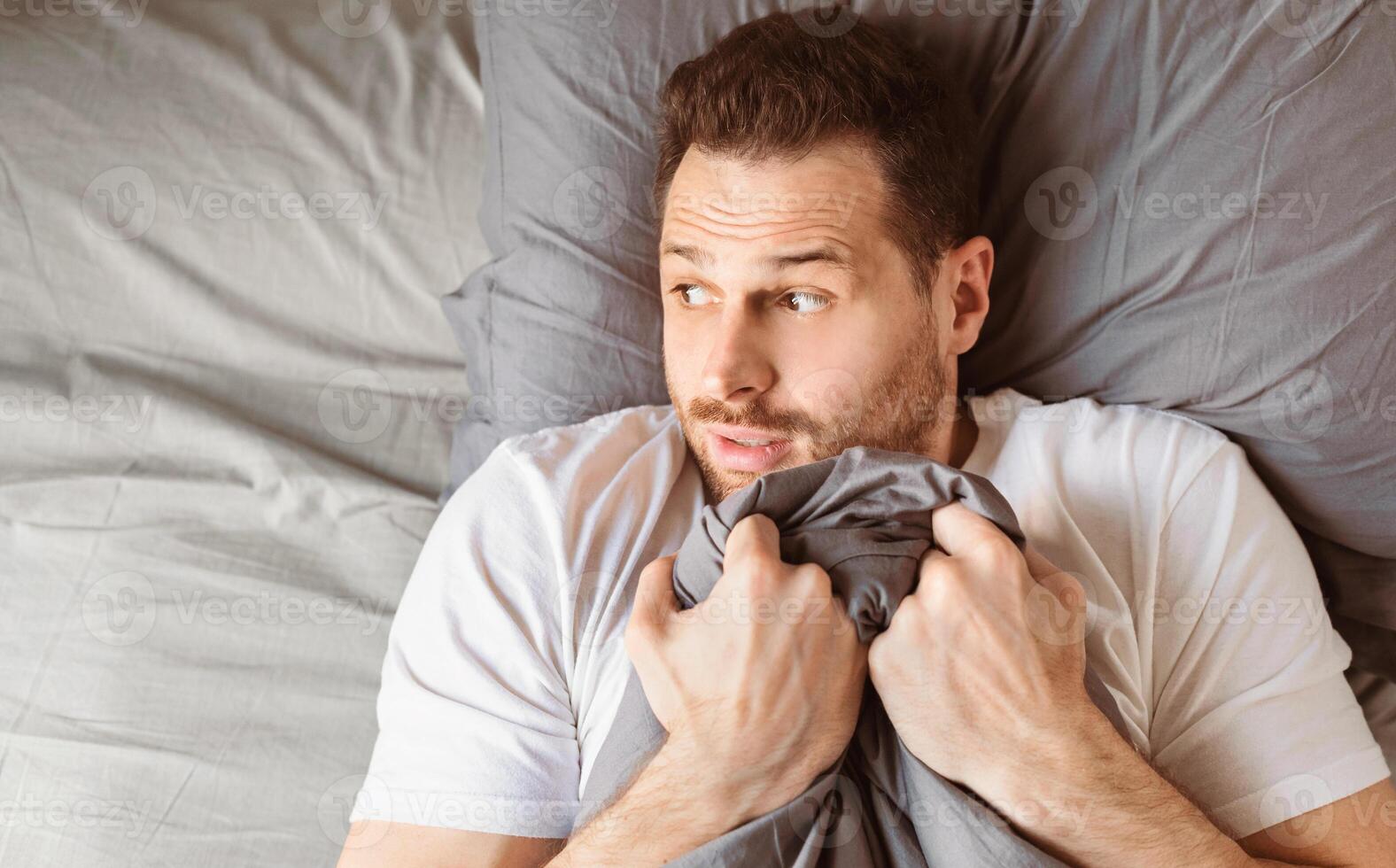 Scared Man Holding Blanket Lying In Bed In Bedroom At Home At Night. Male Panic Attack, Fear And Anxiety. Male Mental Health Problems Concept. Empty Space For Text photo