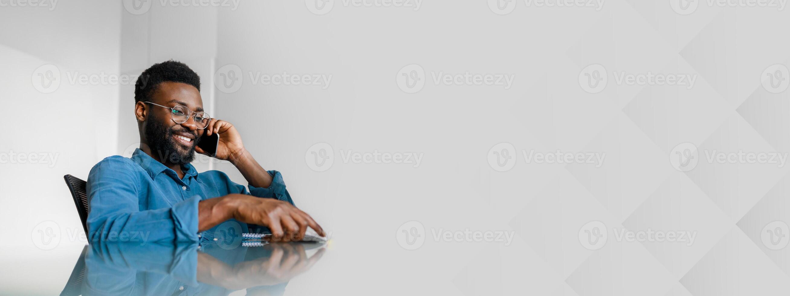 A young African American man, dressed in a blue shirt, engages in a phone conversation while sitting at a sleek office desk. He demonstrates a confident and productive lifestyle with a smile. photo