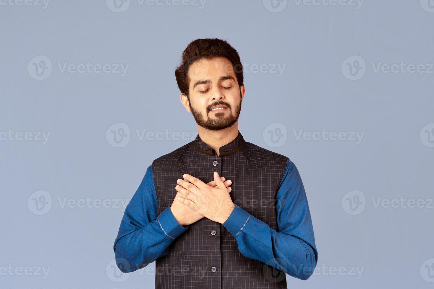 Expressing gratefulness. Portrait of Indian guy with closed eyes holding his hands near heart on lilac background. Panorama photo