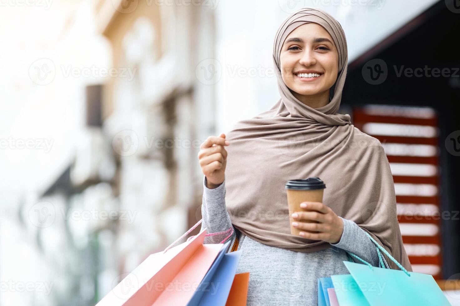 Smiling young woman in hijab holding colorful shopping bags and cup of coffee, copy space photo