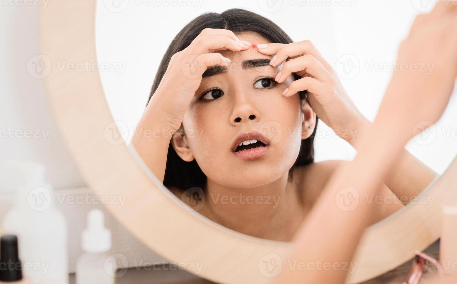 Upset asian young woman checking for pimples, looking at mirror at home, close up, acne and face care concept. Sad lady discovered pimple on her forehead, trying to get rid of it, copy space photo