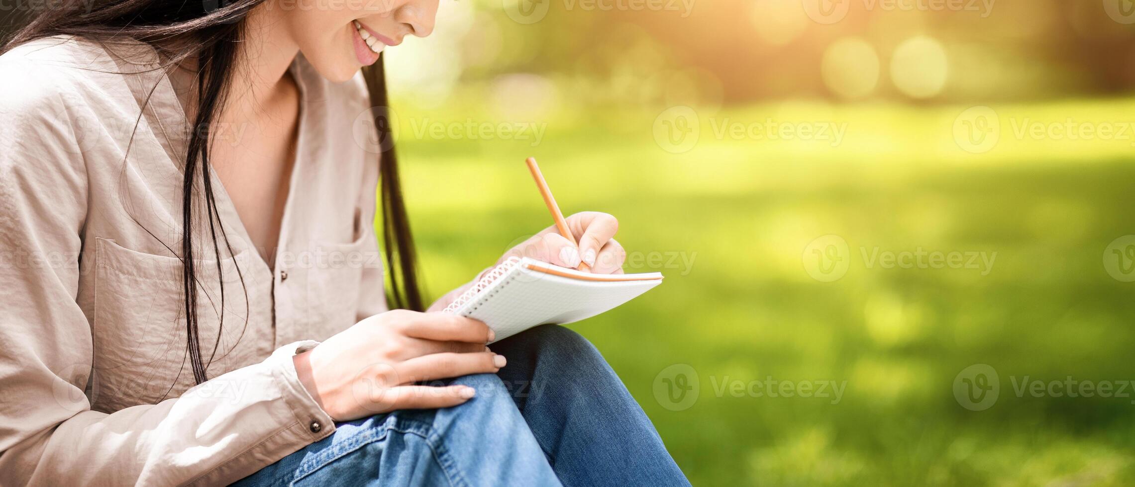 Young asian student girl writing notes to notebook while sitting on lawn in summer park, closeup shot with selective focus and copy space photo