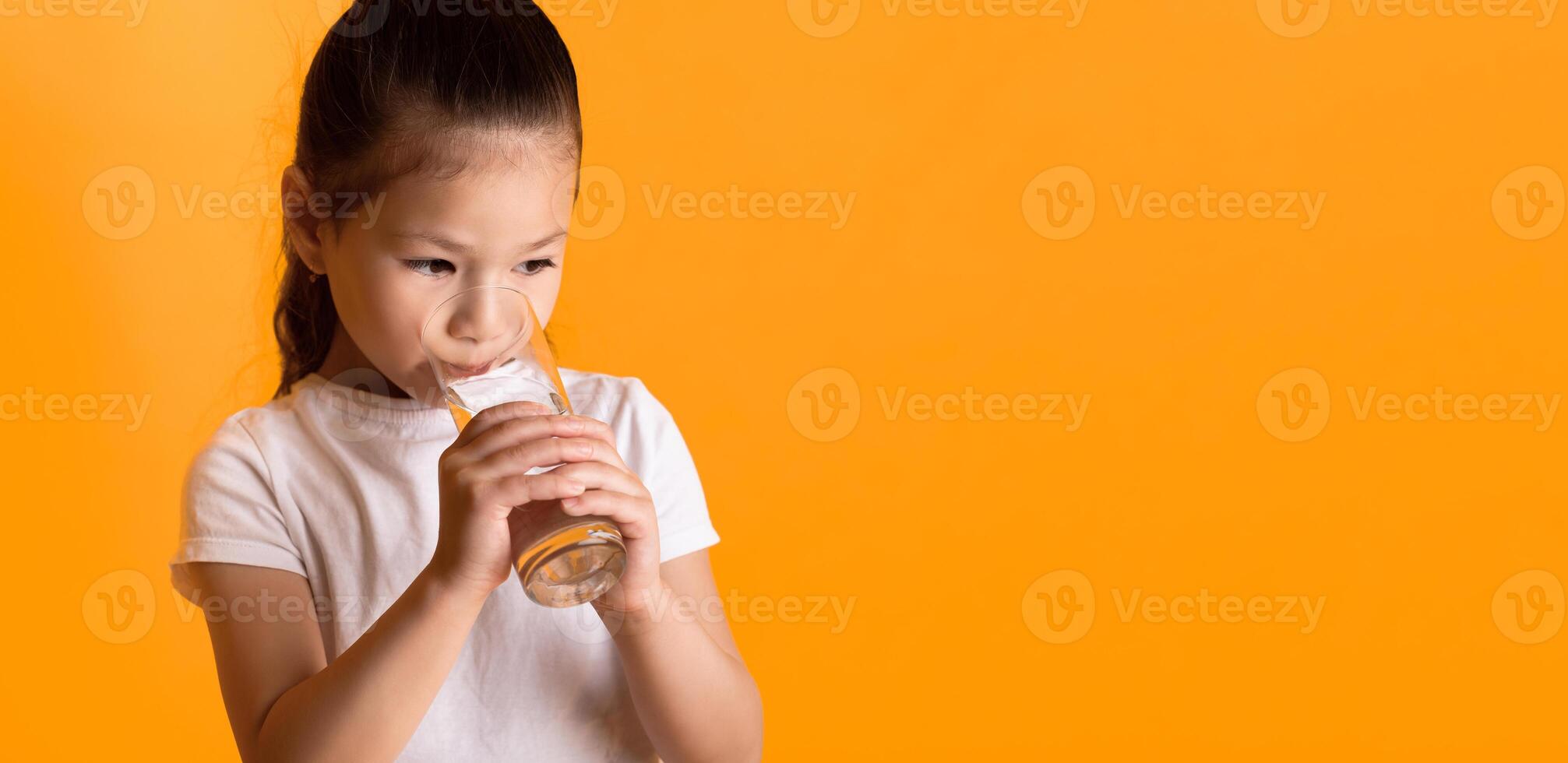 Healthy Beverage. Closeup portrait of thirsty cute asian kid drinking fresh mineral water from glass, looking at copy space. Little girl hydrating her body, isolated over yellow studio background photo